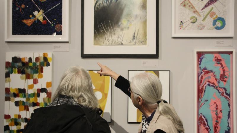 The backs of two women with gray hair side by side. The one on the left is pointing to artwork on the wall.