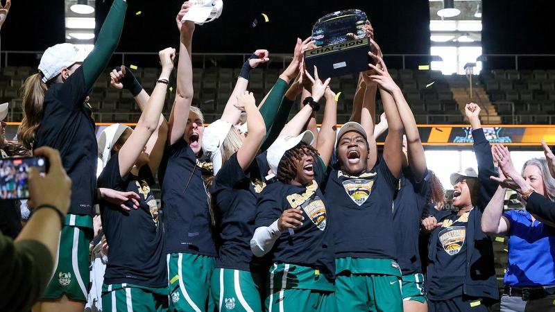 CSU's women's basketball team jump with smiles on their faces holding their 2023 Horizon League Tournament trophy on the basketball court.