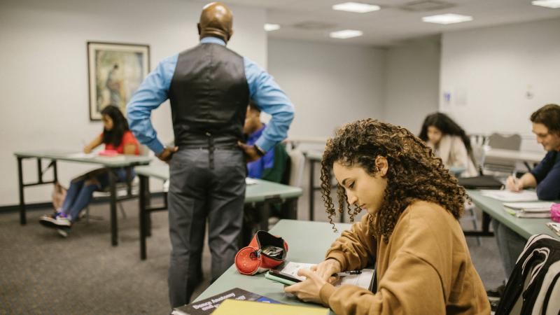 An image of a teacher in a classroom who is turned away from a student texting on their phone.