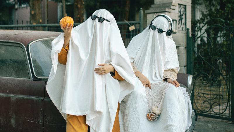 Two people wearing bedsheet ghost costumes pose on the back of a car. 