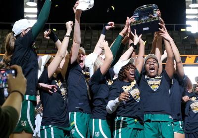 Cleveland State Women's Basketball team jumps with smiles on their faces holding their 2023 Horizon League trophy on the court. 