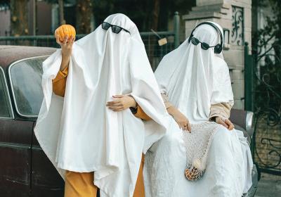 Two people wearing bedsheet ghost costumes pose on the back of a car. 